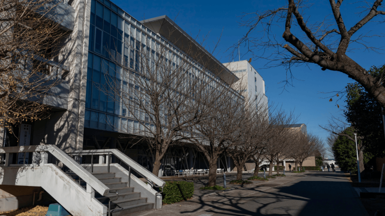 Exterior view of a Nihon University campus building, with a modern glass façade, tree-lined walkway, and students moving through the grounds on a clear day.