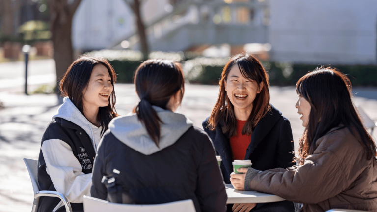 Group of university learners sitting outdoors on campus, chatting and laughing together around a table in a relaxed setting.