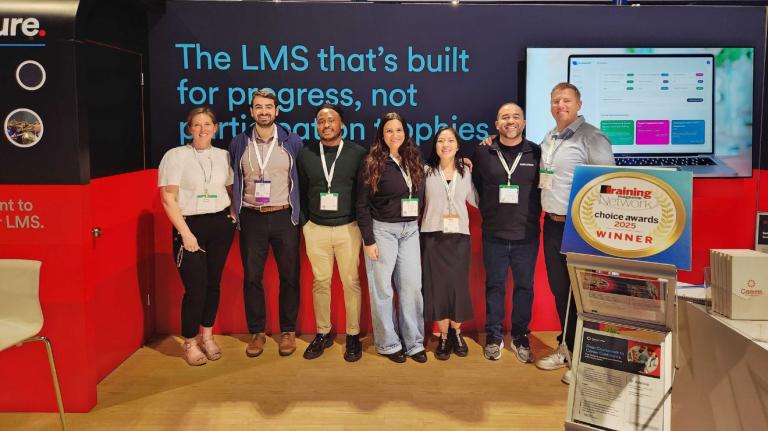 Seven conference attendees standing together at the Instructure Canvas booth during the Training 2026 Conference & Expo in Orlando, with a backdrop that reads ‘The LMS that’s built for progress, not participation trophies.’