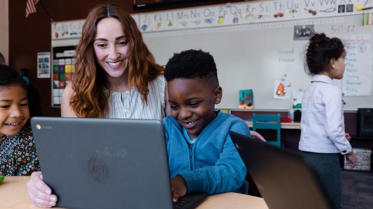 a teacher talks to two students at a table working on a device