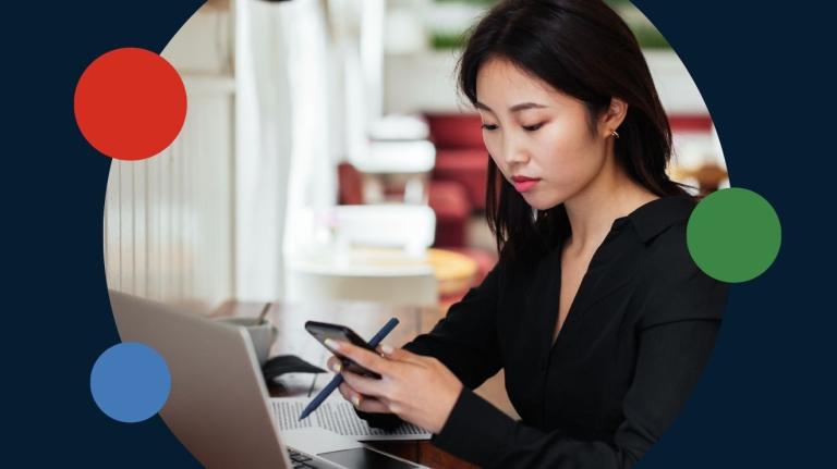 woman with dark hair using a looking at a cell phone at a table with a laptop also in the work area