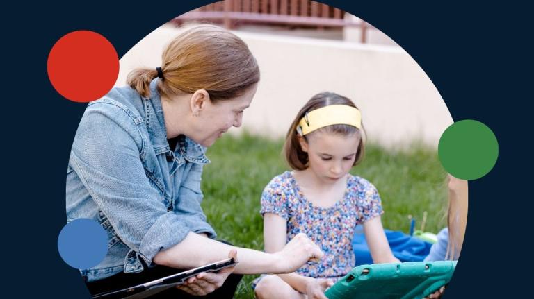 adult instructor sitting on the grass next to a school aged child, working collaborative on tablets