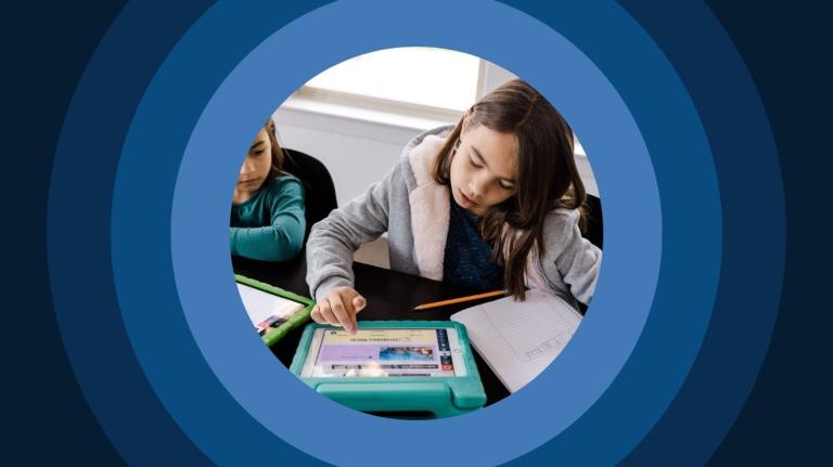 grade school age children sitting next to each other at a table, working on tablet with a nearby notebook
