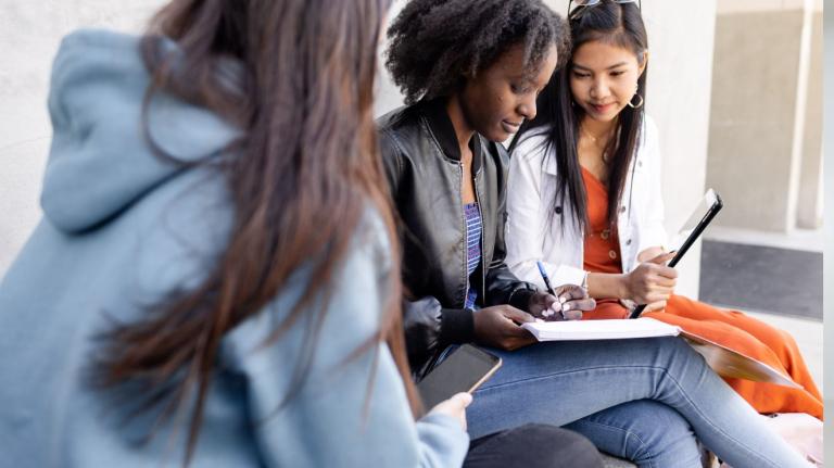 multiracial group of 3 female students, sitting together outside, working on tablets and notebooks
