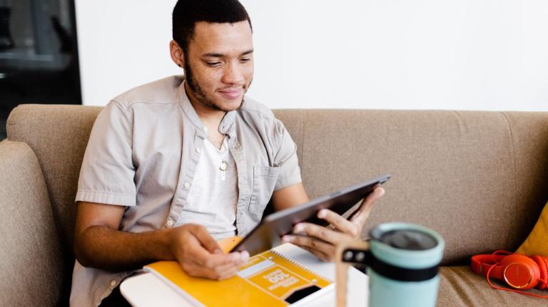 adult male student sitting on a couch working on a tablet, with a notebook, coffee, and headphones nearby 