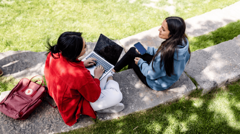 two students talking while using a laptop and a cellphone