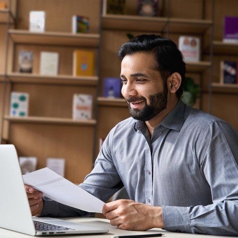 K12 Professional Development - Man in blue collared button down holding paper and smiling at laptop computer