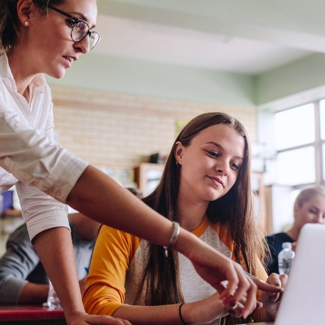 Teacher standing over girl with long hair sitting at classroom desk with laptop