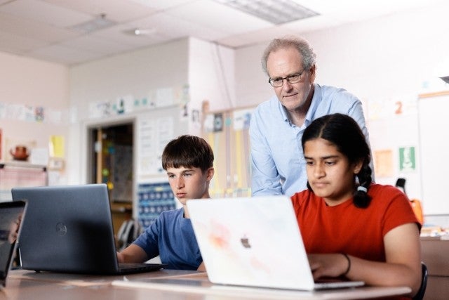 Two students learning on laptops with a teacher in the background.